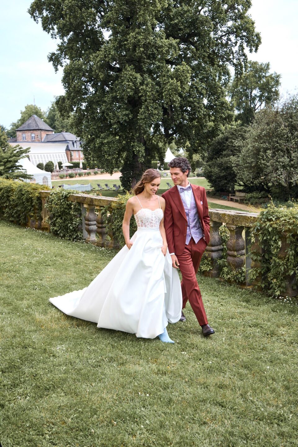 Brautpaar spaziert über die Wiese. Der Brautigam im dunkelroten Hochzeitsanzug lacht seine wunderschöne Braut im langen Satinrock ganz beseelt an während die auf den Boden schaut und dabei lächelt